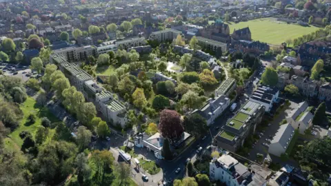 Perkins & Will An aerial computer-generated image of proposed flats and gardens. About half a dozen blocks of flats line one edge of the site with some houses visible on the other side. There are lots of trees and greenery in the middle of the image. Other houses, a section of the Downs green space and sports pitches can be seen around the edge of the site.