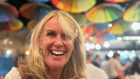 Contributed A picture of Jane Cave. She has a large smile on her face and is wearing a white item of clothing which can be just seen on her shoulders. She has blonde hair and is wearing gold earrings. In the background decorative colourful umbrellas can be seen on the ceiling behind her. 