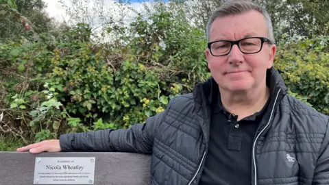 Darren on a bench he had dedicated to his wife. It reads: "Always look for rainbows".