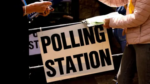 Two people facing one another. We cannot see their heads. The one of the right is wearing a pink coat and is holding a clipboard. In between them is a white sign with the words POLLING STATION written in black.