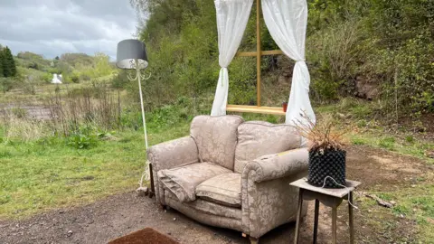 An abandoned beige floral patterned sofa surrounded by a lamp, a side table with a fern-like plant in a black bobbly plant pot, a mat, and a faux wooden window with curtains around it. The living room set up has been dumped on a gravel surface on a patch of land and is surrounded by trees, grass, and a pond.