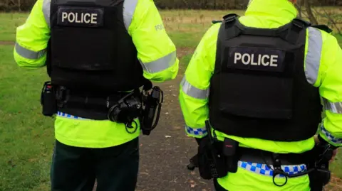 Two PSNI officers in uniform standing in a field with their backs to the camera. Their heads are cropped out of the image.