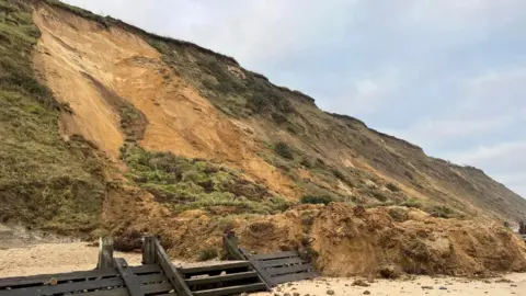 Part of a cliff has fallen away and is on the beach below. Wooden steps and structures can also be seen.