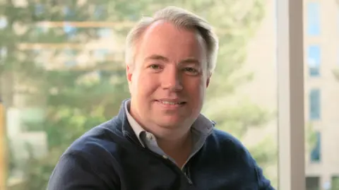 East West Rail Will Gallagher, sitting down, looking at the camera. He is wearing a light blue shirt, a black blue jumper, has great short hair and is smiling. 