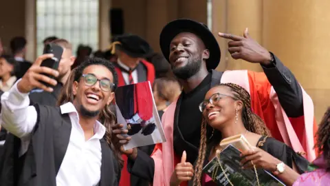 PA Media Stormzy is stood in the middle of two students. The man on the left is holding a phone taking a selfie. They are all looking towards the camera smiling.