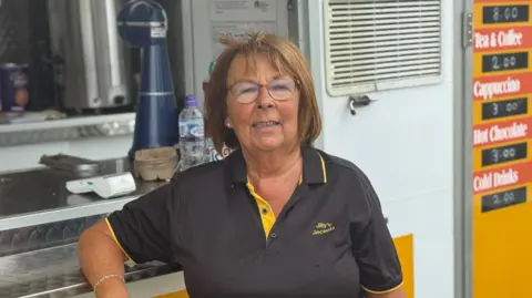 George King/BBC A head and shoulders image of Jill Hales standing in front of her Jilly's Jacket Potatoes stand. She is wearing glasses and smiling into the camera.