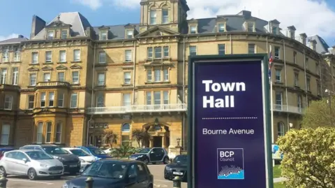 BCP Council A sign says Town Hall and carries the BCP Council logo. Behind it is the five-storey Bournemouth Town Hall, a historical building in light-brown stone with large buildings.