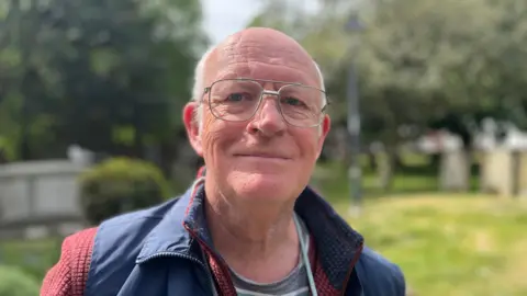 Lucinda Adam/BBC Paul is balding with white hair at the sides. He wears glasses and is dressed in a red jumper and a blue gilet.