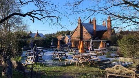 TAG A pub garden scene with a very light dusting of snow seen on the ground among picnic tables and parasols. The garden is deserted with a bright blue sky above.