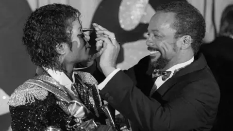 Getty Images Quincy Jones, a man with short black hair and a goatee beard, smiles warmly as he adjusts the sunglasses of Michael Jackson, a younger man who is dressed in an elaborate military-style jacket and has curly black hair. Jones, on the right, is wearing a dinner jacket and bow tie.