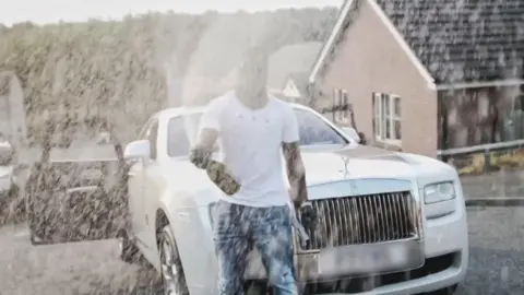 Supplied A man in a white t-shirt sprays champagne in front of a luxury car