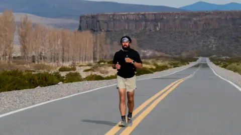 Juan Cruz Lopez A man wearing a black T-shirt and cream shorts in running along a road. In the background is a rocky mountain with bear trees and green bushes to his right.