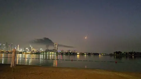 Reuters A view of a city skyline with a missile in the sky that has a trail like a shooting star. The photo is taken from the beach at night and the hotels and high rise buildings are lit up causing reflections in the water.