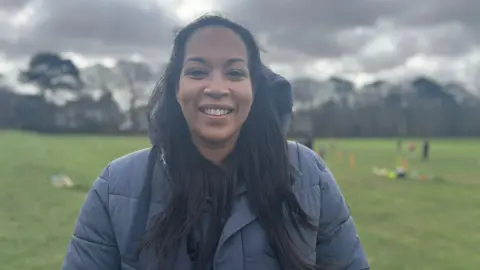 Lauren has long black hair and is wearing a blue jacket. She is smiling at the camera and is stood at the edge of the football pitch.