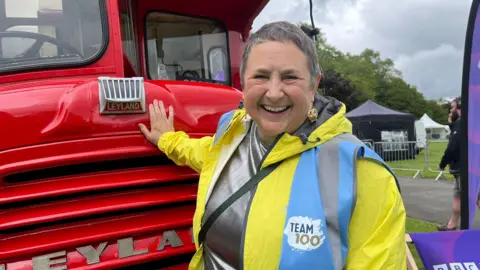 A woman in a bright yellow jacket, with a logo saying Team 100, posing with her hand on a red bus. She has short grey hair and is wearing crown earrings and a silver top. She is smiling widely.