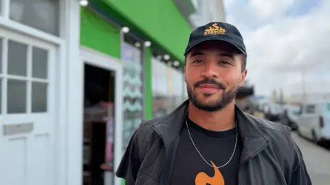 A young man with a beard in a black cap and black t-shirt with an orange flame logo standing in front of a seafront row of shops