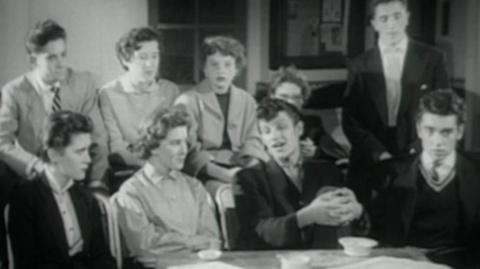 A black and white image of a group of school pupils sat around a table looking like they are being interviewed by someone off mic.  Diane is one of them.