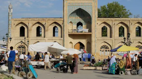 Alamy  Uzbekistan, Ferghana valley, Namangan - people shop at market stalls in front of an old medersa