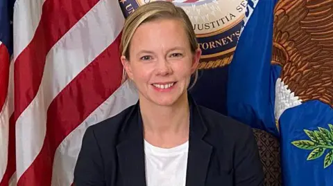 US Department of Justice A woman with short blonde hair looks into the camera with a broad smile. She is wearing a fark jacket and white t-shirt. In the background we can partially see the flag of the United States, the flag of the US Department of Justice and the seal of the US Department of Justice. 