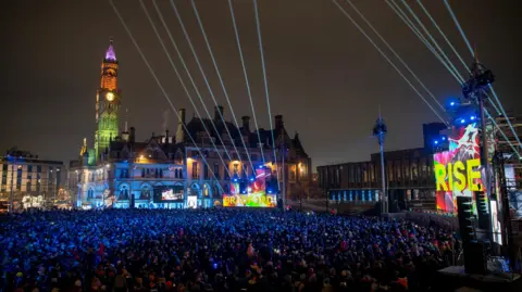 A large open air event in a city square, with a historic town hall and clock tower setting the scene. A large crowd is gathered in front of a stage. There are lots of lasers and neon lights, and big signs saying BRADFORD and RISE.