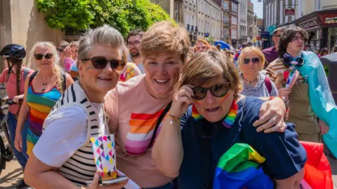 Dani Oliver Three women taking part in a Pride march. There are several people behind them, mostly wearing bright colours.