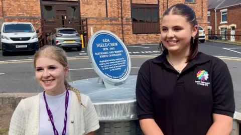 Two females dressed in college uniform stand beside a blue plaque. The girl on the left has blonde hair in a ponytail, white top, purple lanyard and cream cardigan. The girl on the right wears a black polo shirt with a brightly coloured embroidered logo. She has short dark hair. The plaque between the reads 'ADA NIELD CHEW' and stands on a metal plinth. Behind that is a brick building and couple of park cars.