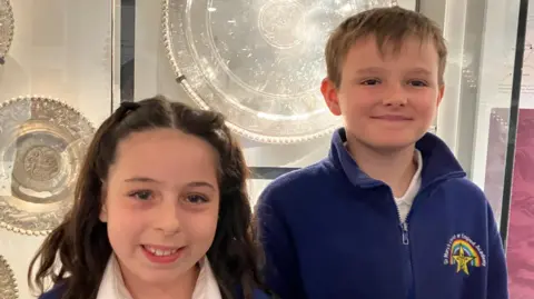 Vikki Irwin/BBC Two young school students - a girl and a boy - smiling while looking into the camera. They are standing in front of a glass cabinet in which there are silvers plates.