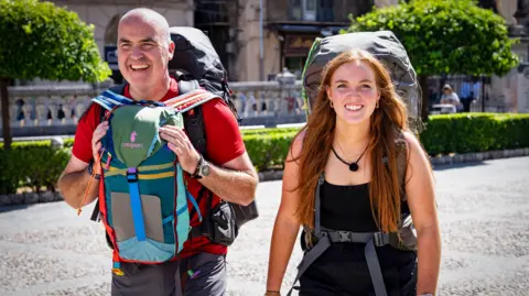 BBC/Studio Lambert Andrew and Molly walking along a street. Andrew is bald, wearing a red t-shirt and carrying rucksacks on his front and back. Molly has long red hair, wearing a black vest and carrying a large rucksack on her back.