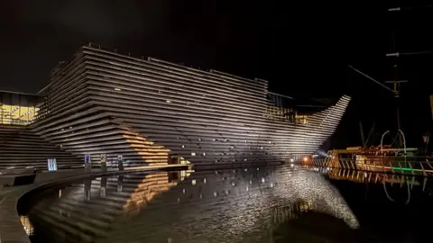 The exterior of the V&A Museum of Design in Dundee taken at night. The building features large concrete sections making up its framework. The museum is reflected in a large pool of water in front of the building 