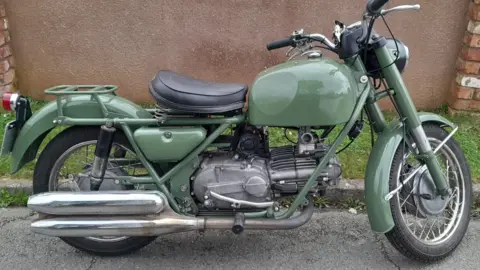 Peter Newton A Moto Guzzi Nuovo Falcone motorbike. It is green. It is parked at a curb. There is green grass and a brown wall. 