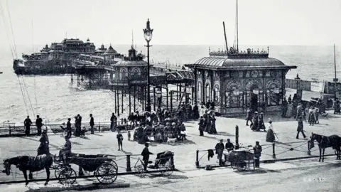 The Regency Society A black and white image of the West Pier. There are horses and carriages in front of the pier, on the road, and groups of people walking. 