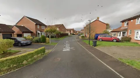 A screengrab taken from Google Maps showing Merlin Park in Portishead. The Image shows a residential street with detached houses, hedges and several cars parked in driveways.