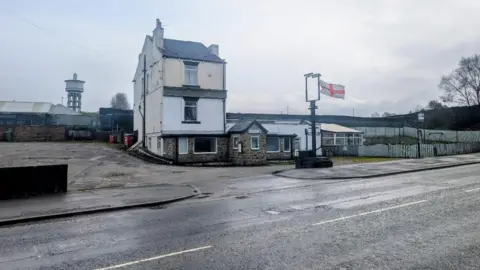 Alex Moss/BBC A three-storey dilapidated building on a roadside. There is a white flag with a red cross on it attached to a metal pole outside. In the background is a water tower. 