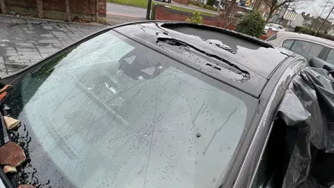 Anand Patel A black car with badly damaged sunroof and cracked windscreen. Black plastic is being used to keep the elements out