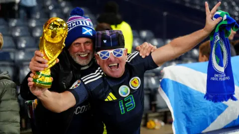 Two smiling Scotland fans holding a replica World Cup trophy at Hampden