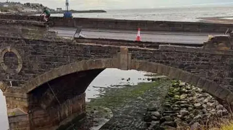 Collapsed section of Banff Bridge. A section of brickwork has falled to the area below. The sea is in the backgroundm with a single orange and white cone on the bridge. 