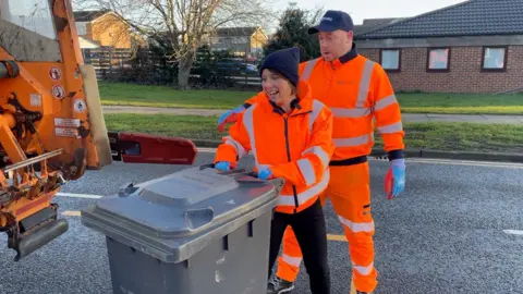 A bin loader and reporter in orange hi-vis manoeuvre a bin onto the back of a wagon.
