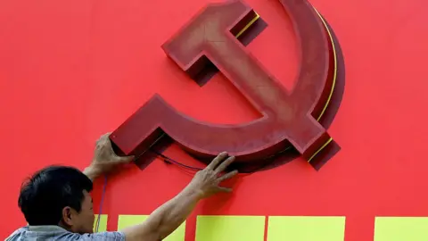 A person reaches up to mount a large cutout of the hammer-and-sickle logo onto a red billboard