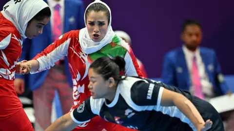 Women kabaddi players dressed in red, white and black. The nearest one, with black hair tied back in a bun, is leaning across the photo while behind her, a woman in red and white with a scarf, tries to reach for her