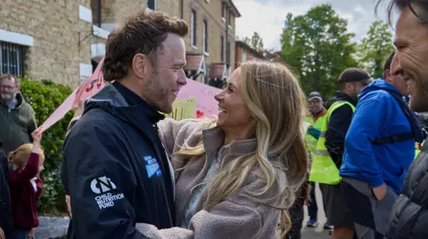 Olly Murs and his wife Amelia look at each other and embrace. There is a crowd around him, some of which are holding signs. Murs is wearing a black sports jacket and Amelia is wearing a brown fleece and has long blonde hair. 