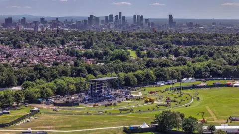 Getty Images Aerial drone view of stage being built at Heaton Park surrounded by construction vehicles in front of rows of trees. In the backdrop are residential houses plus high-rise buildings of Manchester city centre. Pennine hills can be seen in the background.