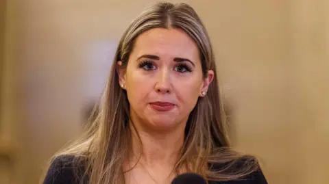 PA Media Sorcha Eastwood has long brown hair and is wearing a black jacket. She is speaking in front of a black microphone.