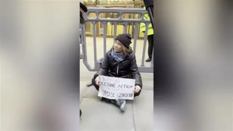 PA Media Greta Thunberg sitting on the pavement behind a temporary barrier, holding a handwritten placard reading “I support Palestine Action prisoners. Oppose genocide,” with police officers nearby.