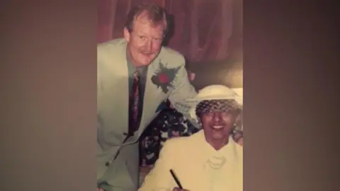 Supplied A photograph of Richard and Chandra on their wedding day. He is wearing a light grey suit with a red rose in his lapel. She is wearing all white including a hat.