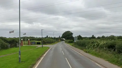 A main road with grass and bushes on each side. A white dotted line leads down the centre of the road and several lamp posts are on the left hand side. Overhead, several wires stream across the sky from cables strung over the road.