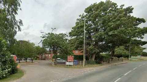 The entrance to Wrangle Primary School. Trees and bushes surround the entrance. The school is off a main A road.