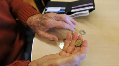 PA Media The hands of an elderly person holding a pound coin at a table that has an open wallet in the background
