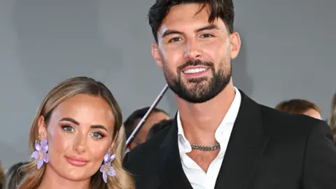 Millie Court and Liam Reardon smiling at the camera, at an event with crowds of people behind them