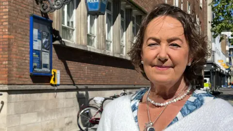 Joanna Brittan is looking at the camera and smiling. She has a short dark bob and is wearing a pearl necklace and earrings. She wears a white fluffy cardigan and is pictured from the shoulders up. She is standing outside a police station.