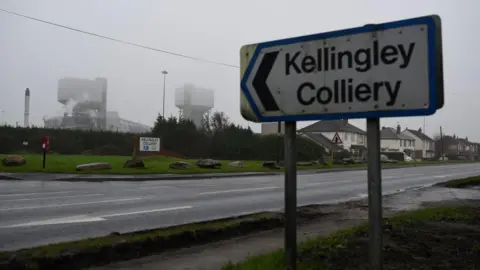 A close up of a road sign in the shape of an arrow which reads 'Keillingley Colliery'. In the background there is a road with industrial buildings to the left ad houses tot he right. 